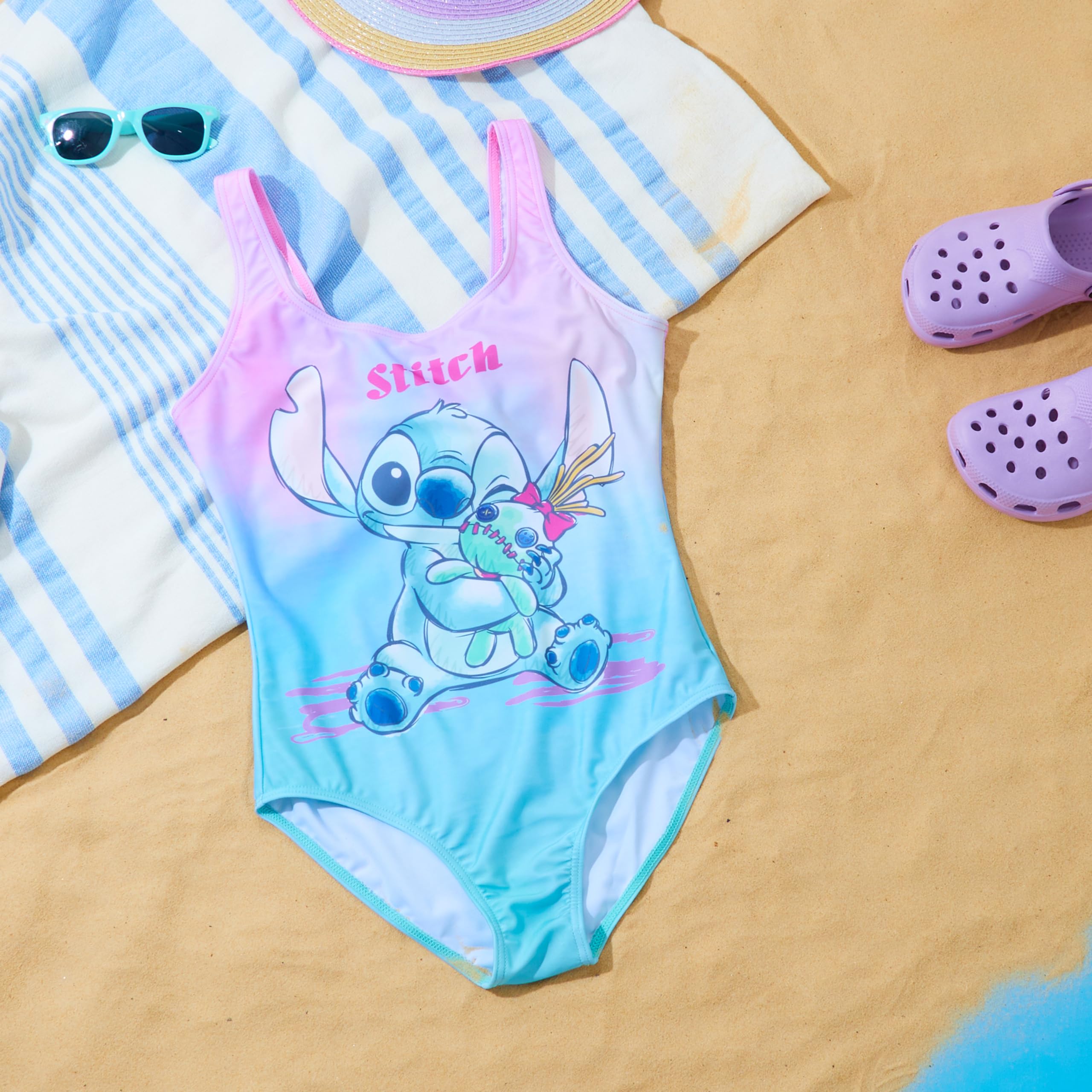 Rosa och blå baddräkt med Stitch-motiv, utlagd på en randig handduk bredvid solglasögon och lila badskor.