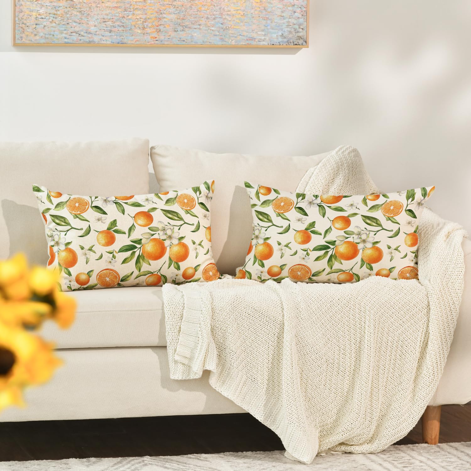 Två kuddfodral med apelsin- och blommönster på en vit soffa med stickad filt.